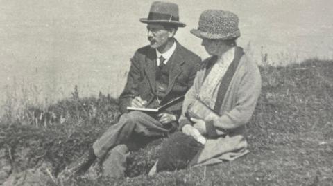 A black and white image of Sydney Bunney and his wife. They are sitting by the coast. He is wearing a hat, dark suit and is sketching in a book. She is also wearing a hat, dress and coat and carrying a handbag. 