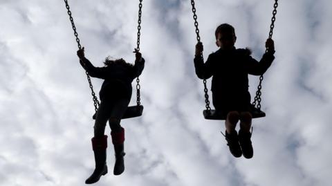 Children on swings. The two children are swinging into the air side by side. Their features are not visible and they appear silhouette-like. It appears to be a young girl on the left and a young boy on the right. The sky behind them is overcast.