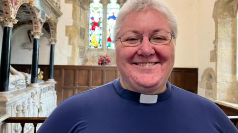 The Reverend Shellie Ward stands inside the church with a stained glass window behind her and an ornamental sculpture to one side. She is wearing blue clerical attire with a collar.