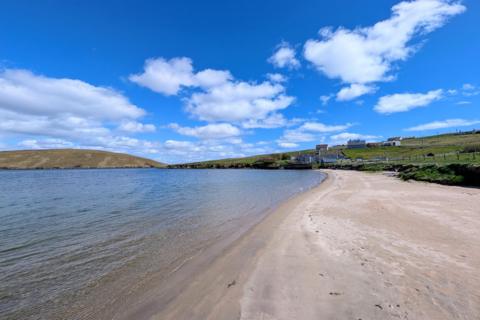 Sandy beach with calm water, grassy hills, small houses, and a bright blue sky in Mid Yell, Shetland Islands