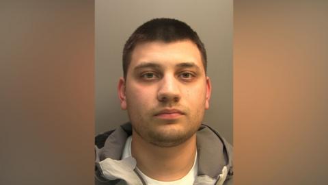 Police custody photograph of Ethan Taylor he has short black hair and beard and is wearing a grey jacket and white shirt