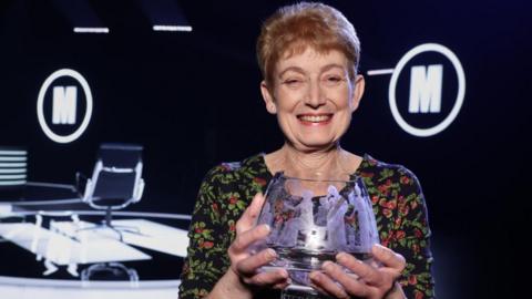 Diane Howe with the Mastermind glass bowl trophy. She has short ginger hair and is wearing a black, green and red flowery dress.