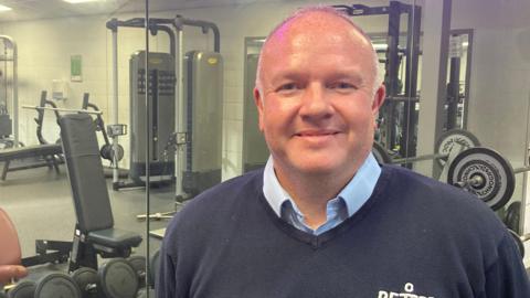 Clive Seabrook standing in a gym inside the Link Centre. He is wearing a light blue collared shirt underneath a navy polo jumper. Behind him is a mirror reflecting the gym equipment and weights on the other side of the room. He has a bald head, is clean shaven and smiling at the camera. 