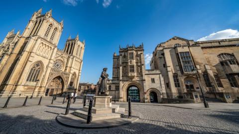 The Bristol Central Library is seen next to the Bristol Cathedral. The sky behind both buildings is blue and there is a roundabout with a cobbled road in front of the buildings.