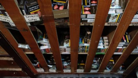 Shelves with boxes of cigarettes on them. Brown wooden bars are in front of the shelves.