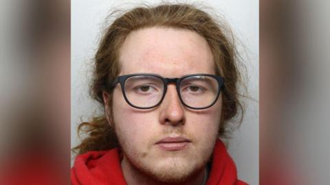 A police mugshot of a sullen man with long, wavy, red hair and black-rimmed glasses. He is wearing a red hoodie.