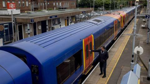 A man pushes the door button on a train on platform two at Sunbury station. The train is in a blue, orange and red South Western Railway livery.