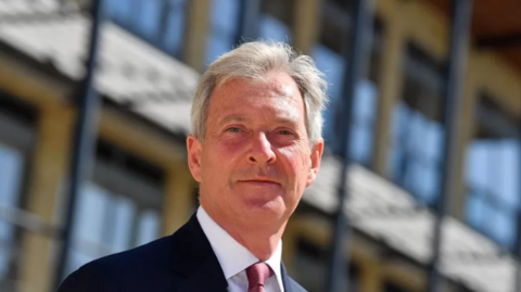 Tim Oliver, wearing a dark suit, white shirt and maroon tie, is pictured standing in front of a modern building on a sunny day.