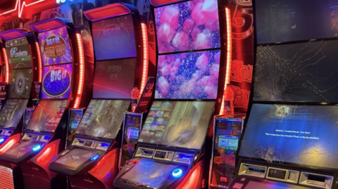Five damaged slot machines with red fluorescent light strips up the sides. All of them have smashed screens.
