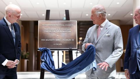 King Charles in a grey suit swiping a cloth off a plaque for the Stephen A Schwarzman Centre for the Humanities.