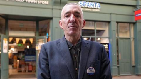 Rob Ellis standing in front of his shop. He has short hair and is wearing a dark blue jacket and shirt