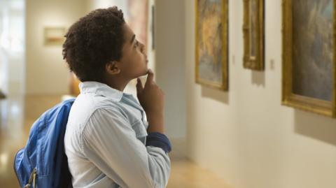 A boy in a light blue shirt and carrying a blue bag looks at a collection of paintings hanging on a wall.