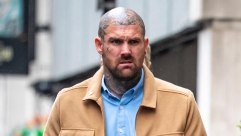 Tony Bartlett walks towards Bristol Crown Court. He is a large man with a shaved head and several tattoos on his neck and head, but not on his face itself. He is wearing a light blue shirt under a light brown jacket.