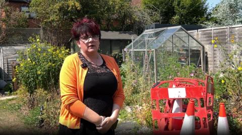 A woman stood next to some red cones and barriers in her garden next to some plants and a green house