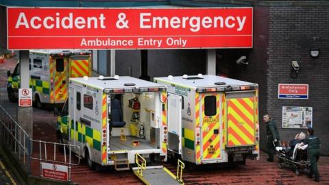 Two ambulances parked outside an A&E department. One has its rear doors closed while the other has its doors open. A paramedic, wearing green overalls, can be seen adjusting a bed in the rear of the vehicle.