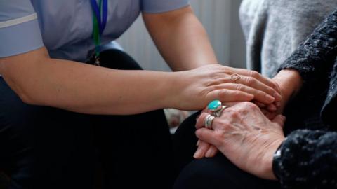 A carer has her hands placed on an older woman's hands. They are both seated and in a room.