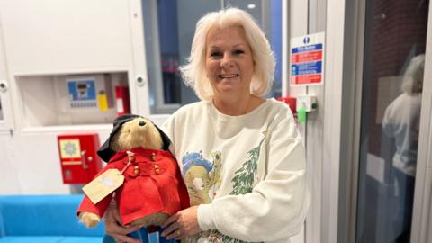 A woman in a white jumper with a bear and a Christmas tree on holds a Paddington Bear wearing a red jacket and black hat. 