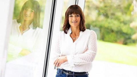 Image shows Dani Barnett - smiling, with medium-length brown hair. She wears a white blouse with jeans, silver necklace and is leaning against a window with a garden in the background, her reflection visible.