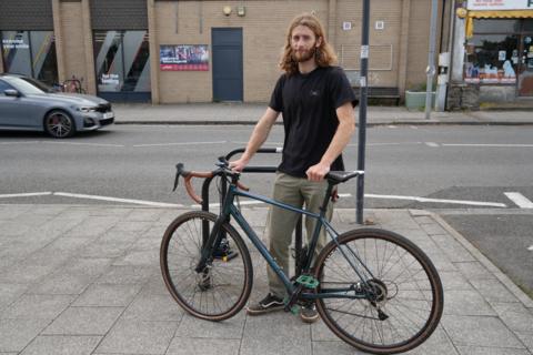 Josh is standing on the pavement holding his bike 