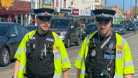 Two police officers walking in front of a parade of shops in Southend-on-Sea on a sunny day.