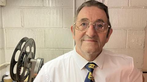Pat Church wearing a white shirt and tie with the word FILM written on it. He is wearing glasses and looking into the camera. A reel projector is on the left of his shoulder. 
