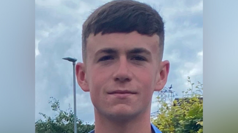 A young man looks at the camera with a slight smile. He has brown hair that falls over his forehead. A lamppost and hedges are in the background.