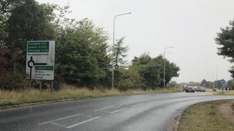Part of the A12 through Suffolk. It shows a road leading up to a roundabout. Cars can be seen in the distance making use of the roundabout. There is a road sign and trees and bushes line the road.