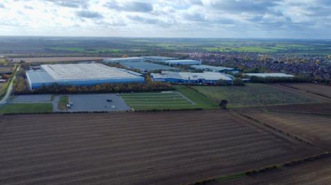 An aerial view of Thrapston and the current industrial estate, which consists of several very large white or pale blue warehouses. In the foreground are brown fields and in the background, green fields with hedgerows.