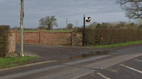 The entrance to what appears to be a farm drive. There is a hedge alongside and a road with white road marking in front. There is also a sign post and a telegraph pole on either side of the entrance.