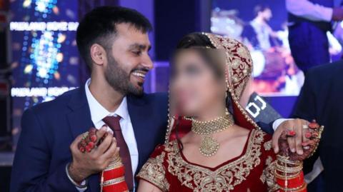 Jagtar Singh Johal wears a navy suit with a dark red tie and white shirt and holds Gurpreet Kaur's hands as they dance. She wears a red sari with gold embroidery and a gold necklace.