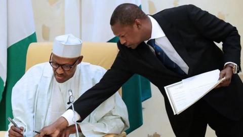 Nigeria’s former justice minister Abubakar Malami in a black suits lean towards seated former President Muhammadu Buhari dressed in a white gown and cap, showing him something on a desk