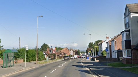 A road with houses on both sides and a pedestrian crossing with traffic lights. There is a green bus stop on the left-hand side pavement. There are cars driving down the road.