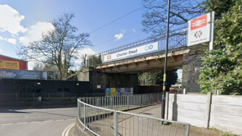 A road with metal railings in the foreground and a bridge overhead with the words Chester Road on it