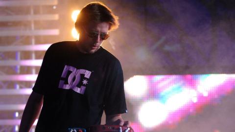 Groove Armada member Andy Cato on stage at a festival. He has a black top with a white logo on it and there are large neon lights behind him. He is wearing sunglasses