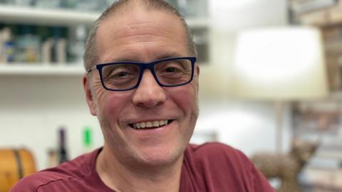 A man smiles as he sits in a home in front of a lamp and a book shelf, which are out of focus. He wears glasses with dark-blue frames and a dark-red t-shirt.