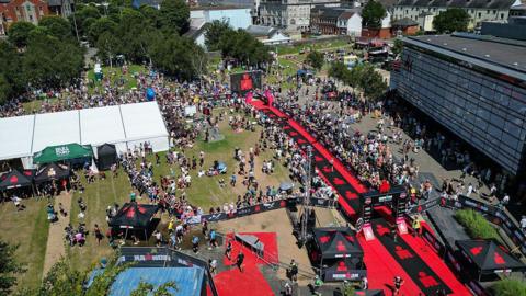 A bird's eye view of an event space, with large crowds of people gathered. On the right of the picture is a large building, and on the left is a white tent. A red runway can be seen running through the middle of the picture. 
