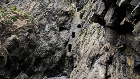 Wide view looking up through a rocky gorge with jagged grey cliff walls on both sides. A tall, narrow stone structure is embedded between the cliffs, with small window openings, and a dry, pebble-covered riverbed lies at the bottom.