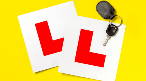 Two bright white square cards sit against a bold yellow backdrop, each marked with a large red “L” — the unmistakable symbol of a learner driver. Beside them lies a set of car keys with a black fob