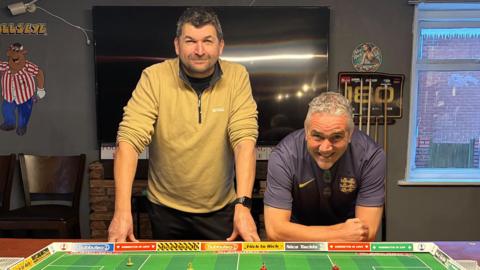 Steve Race is standing on the left and is standing upright in front of a table with a Subbuteo set on it. He is wearing black trousers with a quarter zip jumper and is looking directly at the camera and smiling. Next to him on the right is club member Adam Neckrews who is leaning over in front of the table and is wearing a purple football shirt. He is also looking directly at the camera and smiling.