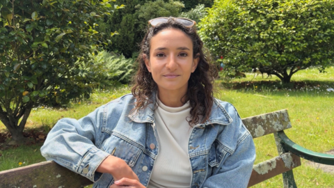 Young woman with brown curly hair, sat on a park bench, wearing a blue denim jacket. 