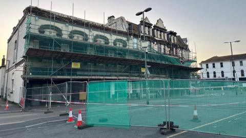 The detached building is four storeys high and the front of it is covered in scaffolding which is netted with green mesh. In front of the building is high metal fencing, some of which is also covered in green mesh, some traffic cones and some red and white tape.