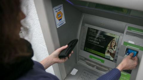 Close-up of a woman using an outdoor ATM machine. The person is holding a wallet in one hand and inserting a blue bank card with the other. The ATM is grey with green accents around the card slot and keypad and the screen displays a welcome message. A security sticker reading ‘Secured by ADT’ is visible on the side panel.