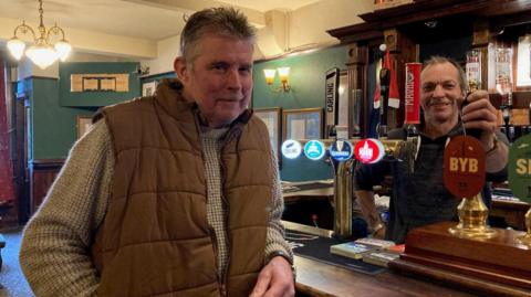 A man wearing a brown gilet and mottled grey jumper leaning on a brown wooden bar with a second man behind the bar holding on to a brass-topped, wooden handled beer pump.