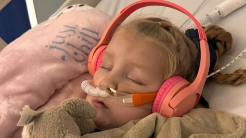 Graycen is asleep in a hospital bed with pink headphones with hospital tubes in her nose. There's a couple of blankets on top of her and a teddy bear.