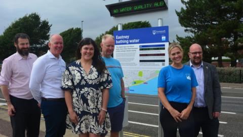 Various councillors and local activists stand in front of a blue sign titled 'Eastney bathing water' with a road behind them. Above the sign, there's an electronic display saying 'no water quality alert' in green letters.
