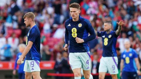 Scotland player Scott McTominay. He is wearing a dark blue Scotland branded quarter zip top while mid jog. Fellow players can be seen in the background.