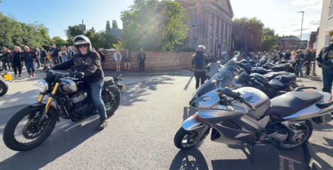 To the right of the photo are a row of motorbikes all lined up on the road. People are gathered around to look at them. To their right is a man who is riding a black motorbike. He is wearing a black jacket and black helmet. 