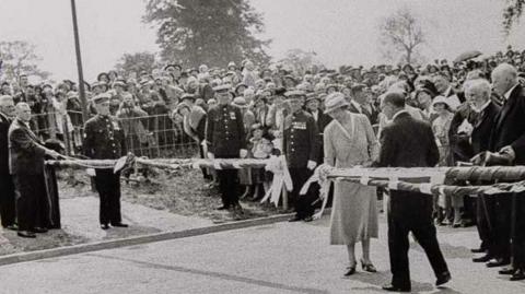 A black and white photo of a woman in light dress and hat, cutting a ribbon on road barriers