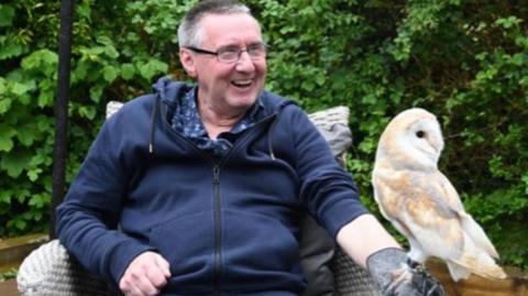 A middle aged man sits wearing a blue hoodie with a large grey glove on his left hand. A white owl sits on the glove.