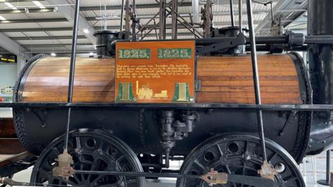 Locomotion No 1 on display in a museum. It has large black wheels and is the shape of a large barrel, the top half of which is made of wooden slats. The name plates are displayed on a large wooden board on its side.